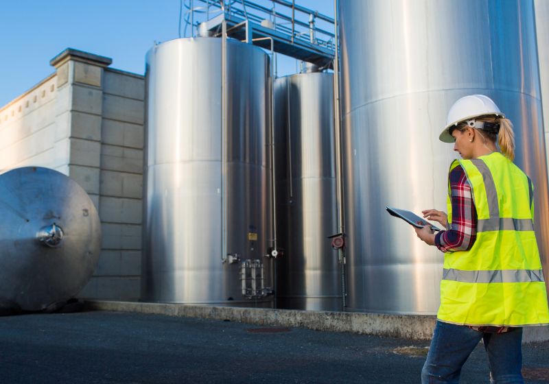 A person using a tablet wearing a hardhat and yellow vest in front of large silver liquid tanks.