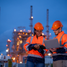 Two people in hardhats and vests look at a computer in in front of a factory.