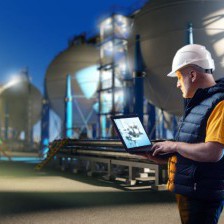 A person holding a laptop and wearing a hardhat at night surrounded by large spherical tanks.